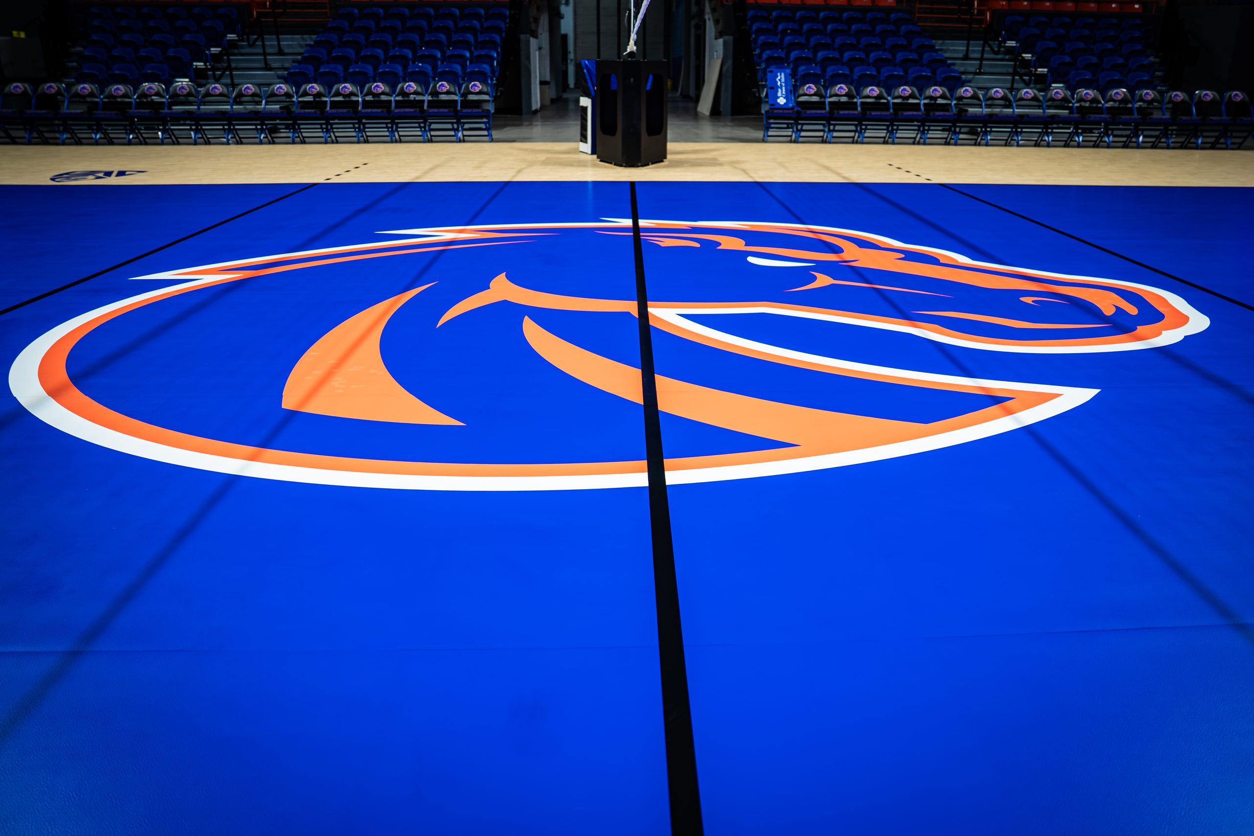 A vibrant blue basketball court features a large orange and white horse logo at center, with empty bleachers in the background.