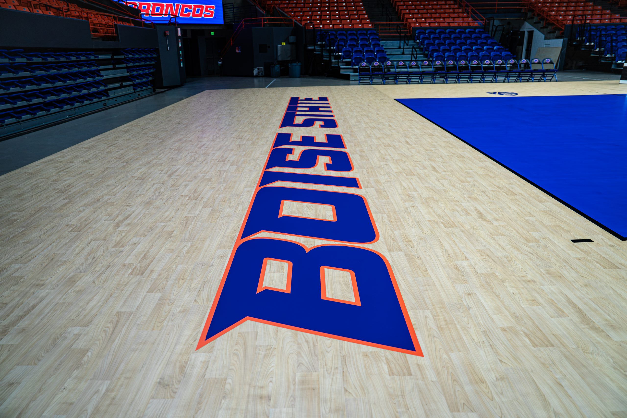 A basketball court with "BOISE STATE" in bold blue and orange letters, surrounded by empty blue seats and a digital scoreboard displaying "BRONCOS".