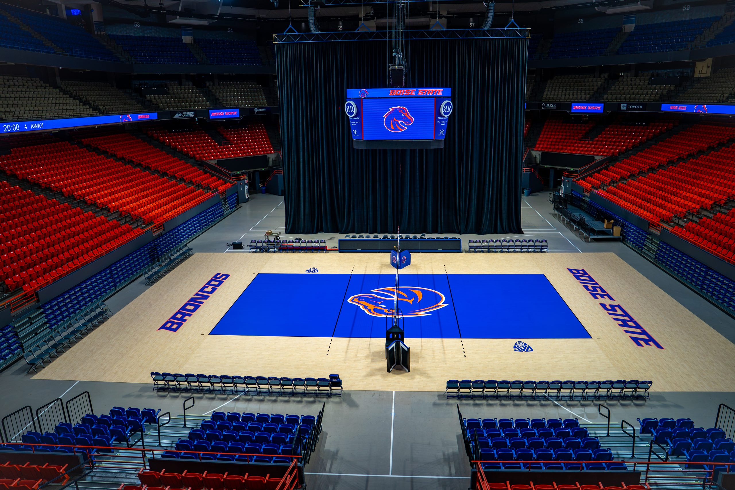 An empty indoor basketball arena with a blue court featuring the "Broncos" logo and "BYU STATE" text, surrounded by red and blue seating.